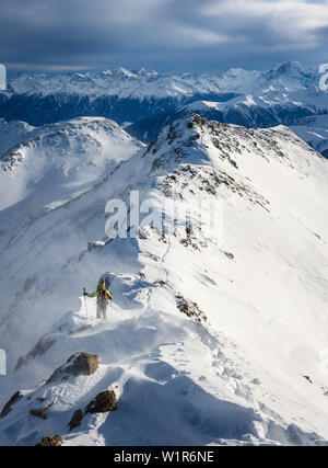 Young ski mountaineer travereses on a snowy ridge with skis on the backpack in windy conditions, Sesvenna, Vinschgau, South Tyrol, Italy Stock Photo