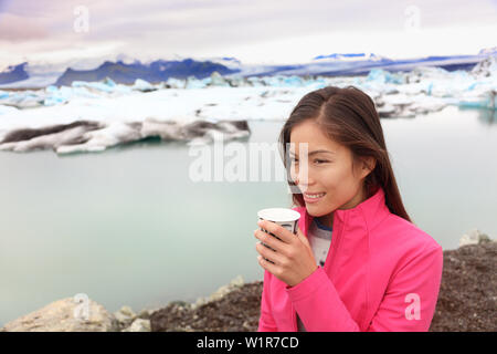 Woman drinking coffee on travel trip at glacier lagoon on Iceland. Happy tourist woman enjoying view of Jokulsarlon glacial lake. Smiling woman in beautiful Icelandic nature landscape with icebergs. Stock Photo