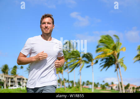 Healthy active man runner running in tropical park. Portrait of handsome young male jogger training cardio going for a run in city park or resort with palm trees in the background in summer. Stock Photo