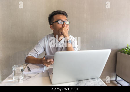Freelancer with cellphone , computer putting hand on chin while thinking Stock Photo