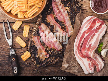 Bacon flavored snacks chips with grilled bacon rashers on vintage chopping board with smoked raw bacon on butchers paper with garlic and sauce on wood Stock Photo
