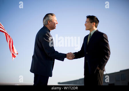 Confident young adult businessman shaking hands with a mature male colleague with a flag behind. Stock Photo