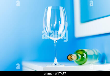 Empty wine glass on a white table next to a lying bottle of white wine on a blue background Stock Photo