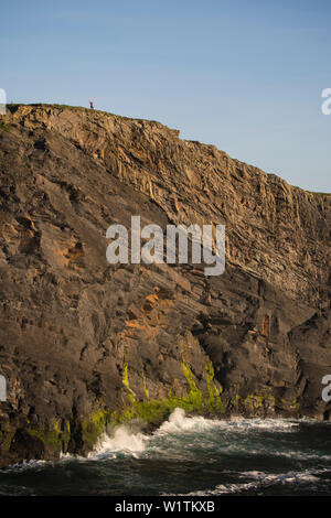 A seemingly tiny figure - a woman - stretches her arms on top of the Kilkee Cliffs and thereby puts the height in relation, Kilkee, County Clare, Irel Stock Photo
