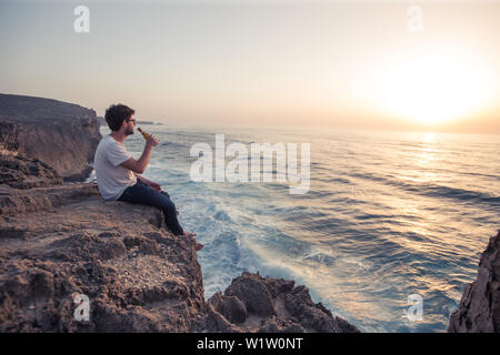Young man sitting on a cliff edge at the sea at the beach Praia da Amoreira, Aljezur, Faro, Portugal Stock Photo