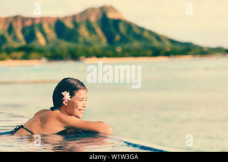 Smiling beautiful woman relaxing in infinity swimming pool. Side view of happy female looking at ocean view on sunny day. Young tourist is enjoying summer vacation against mountain. Stock Photo