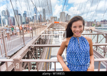 Beautiful young Asian woman portrait on Brooklyn bridge, New York city NYC, Manhattan, USA. Smiling tourist in blue dress doing summer travel in urban landmark.