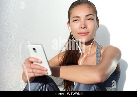 Young woman listening to music with earphones on smart phone app for fitness motivation. Athlete runner in sportswear relaxing sitting getting inspired. Asian mixed race model. Stock Photo