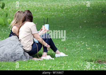 Two girls sitting on a green grass, drinking cocktails and sharing impressions. Summer leisure on a clover meadow, concept of female friendship Stock Photo