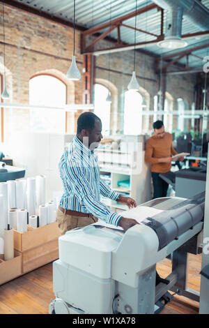 In printing office. Dark-skinned man wearing striped shirt working in printing office waiting for the poster Stock Photo