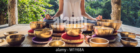 Woman playing on Tibetan singing bowl while sitting on yoga mat against a waterfall. Vintage tonned. Beautiful girl with mala beads meditating BANNER Stock Photo