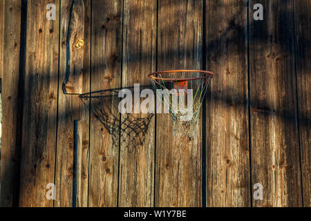an old shabby basketball hoop is throwing an evening shadow on a weathered wooden wall Stock Photo