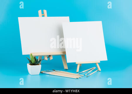 Two wooden blank easels with brushes and plant on blue background. Stock Photo