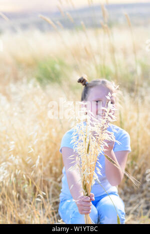 Beautiful girl holding spikes of wheat and ears of oats. Cute child sitting on the gold summer field ready for harvest. Selective focus on spikes. Stock Photo