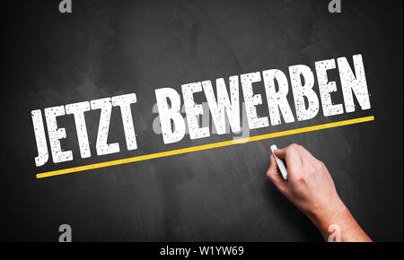 hand writing 'Apply Now!' in German on a chalkboard Stock Photo