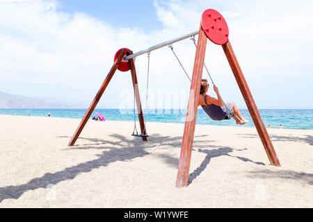 middle aged woman swinging on a swing. On the beach with the sun on her face in Spain. Stock Photo