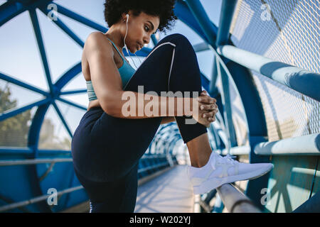 Fitness woman doing stretching workout of the legs on a bridge railing. African woman doing workout on a walkway bridge in morning. Stock Photo