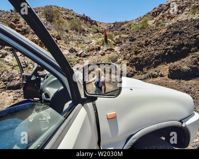 Partial view of a big car, in the right side mirror is a woman to see in the background the barren desert landscape at the foot of the Teide in Teneri