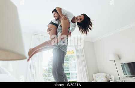 Young man standing on bed and carrying his girlfriend on his shoulder. Couple in playful mood in bedroom. Stock Photo