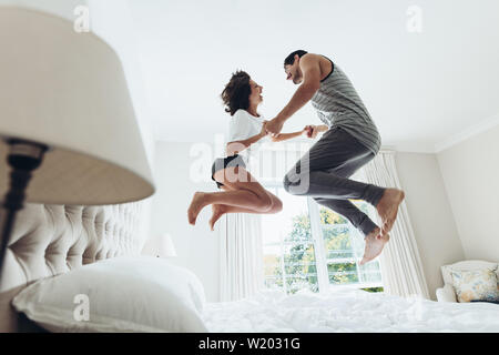 Man and woman holding hands and jumping together on bed. Couple having fun in bedroom. Stock Photo