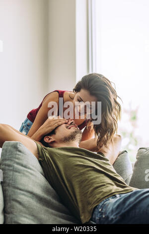 Young woman surprising man sitting in the living room at home. Smiling female covering eyes of her boyfriend. Stock Photo