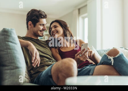 Loving couple sitting on sofa at home. Man and woman relaxing on couch with smartphone in living room. Stock Photo