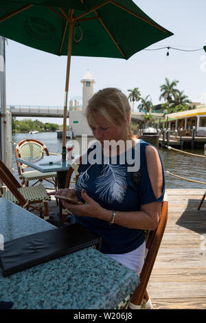A woman checks her phone while sitting at a table under an umbrella on the deck at Two Georges Waterfront Grille in Boynton Beach, Florida, USA. Stock Photo