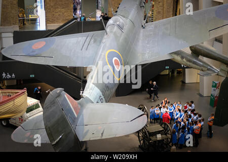 School class gathers below World War II Spitfire to hear about the war, London. Stock Photo