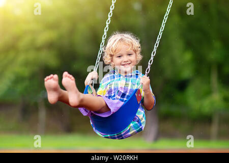 Child playing on outdoor playground in rain. Kids play on school or kindergarten yard. Active kid on colorful swing. Healthy summer activity for child Stock Photo