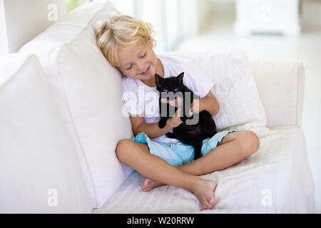 Child playing with baby cat. Kid holding black kitten. Little boy snuggling cute pet animal sitting on white couch in sunny living room at home. Kids Stock Photo