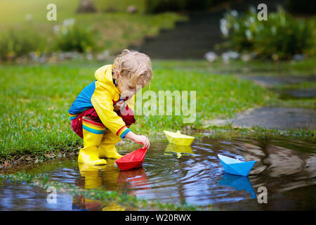 Child playing with paper boat in puddle. Kids play outdoor by autumn rain. Fall rainy weather outdoors activity for young children. Kid jumping in mud Stock Photo