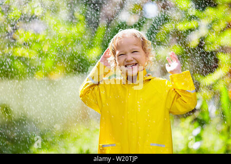 Child playing in the rain on sunny autumn day. Kid under heavy shower with yellow duck umbrella. Little boy with duckling waterproof shoes. Rubber wel Stock Photo