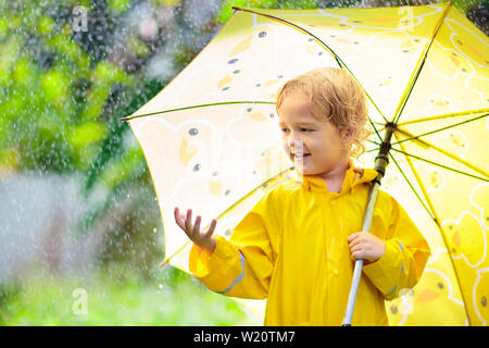 Child playing in the rain on sunny autumn day. Kid under heavy shower with yellow duck umbrella. Little boy with duckling waterproof shoes. Rubber wel Stock Photo