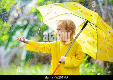 Child playing in the rain on sunny autumn day. Kid under heavy shower with yellow duck umbrella. Little boy with duckling waterproof shoes. Rubber wel Stock Photo