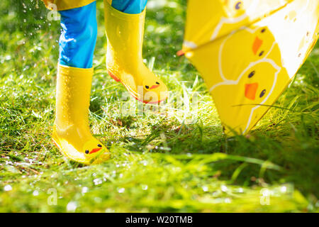 Child playing in the rain on sunny autumn day. Kid under heavy shower with yellow duck umbrella. Little boy with duckling waterproof shoes. Rubber wel Stock Photo