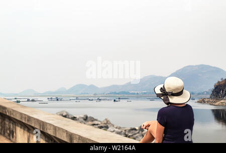 Woman sitting on concrete slab Background The raft floating fish farming in water and mountains at Krasiew dam , Supanburi Thailand Stock Photo