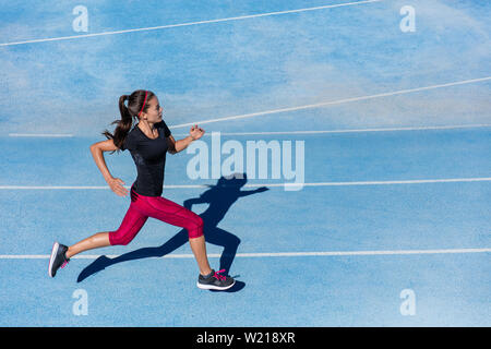 Athlete runner running on athletic track training her cardio. Jogger woman jogging at fast pace for competition race on blue lane at summer outdoor stadium wearing red capri tights and sports shoes. Stock Photo