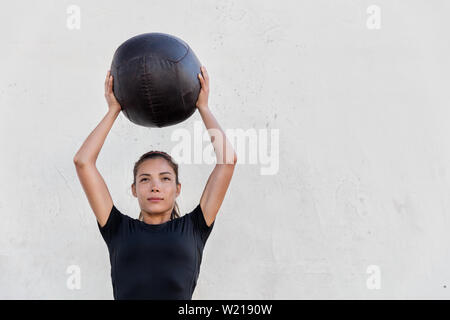 Fitness crossfit girl holding medicine ball above head for shoulder press workout in outdoor crossfit gym. Young Asian athlete girl doing upper body exercise working out with heavy weighted balls. Stock Photo