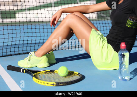 Lower body closeup of tennis player woman resting sitting on outdoor court showing racquet / tennis racket, ball and water bottle wearing yellow sportswear outfit. Sports shoes and skirt. Stock Photo