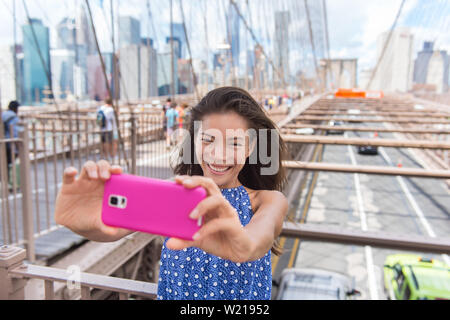 Happy New York City selfie tourist young woman taking a self-portrait photo with smartphone app on Brooklyn Bridge, NYC, Manhattan, USA. Asian girl doing mobile phone photography for social media. Stock Photo