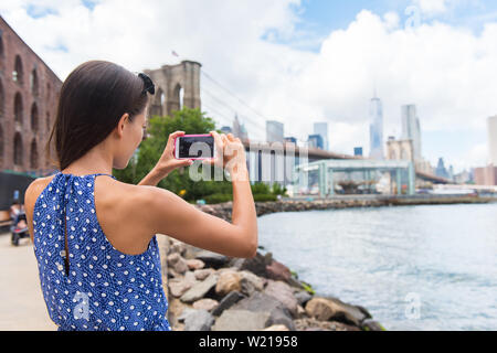 Tourist taking travel picture with phone of Brooklyn bridge and New York City skyline during summer holidays. Unrecognizable female young adult enjoying USA vacations in blue dress. Stock Photo