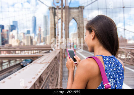 Smartphone texting girl on Brooklyn bridge in urban New York City, Manhattan USA. View from the back of unrecognizable business woman holding phone reading or using social media in summer. Stock Photo