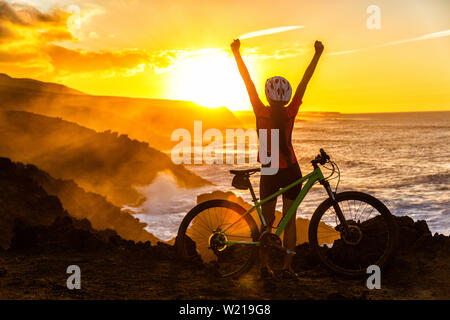 Success, achievement, accomplishment and winning concept with cyclist mountain biking. Winning happy MTB woman cycling reaching goal raising arms at sunset cheering and celebrating at summit top. Stock Photo