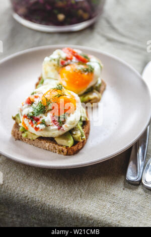 Breakfast served of two toasts with avocado, fried eggs with vegetables and herbs on a rustic tablecloth background. View from above. Healthy and nutr Stock Photo
