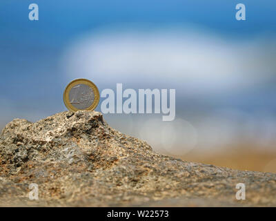 One Euro coin on rocky beach against blurred blue sea Stock Photo