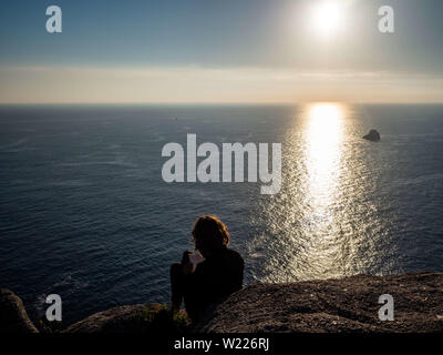 June 27, 2019 - Finisterra, A CoruÃ±a, Spain - A woman is seen writing on a rock while waiting for the sunset. Cape Finisterre is the destination of those pilgrims who, after visiting the St Jamesâ€™ tomb is Santiago, continue their way along the route marked out for them overhead by the Milky Way until they could go no further. Finisterre was considered during the period of Classical Antiquity to be the end of the known world. Finisterre, or Fisterra in Gallego, the local Galician dialect, means ''Land's End'' in Latin. Next, to the lighthouse,  there is the 0.0 Kilometer Marker from where th Stock Photo