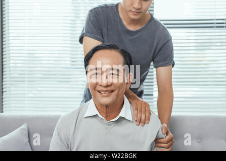 Asian old man shoulder pain, sitting on sofa , son massaging father shoulder. Stock Photo