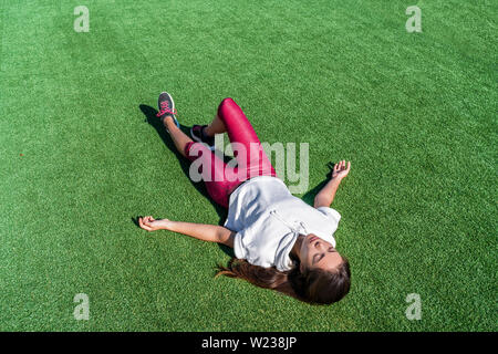 Tired athlete lying down from heat exhaustion on grass park after intense workout. Exhausted Asian runner woman taking a break sweating during cardio running exercise training in summer outdoors. Stock Photo