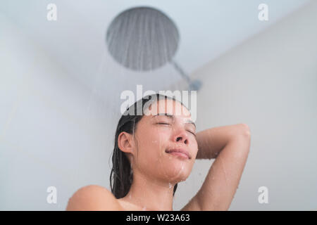 Young woman taking a hot shower showering hair relaxing under warm running water from modern rainfall shower head at home or hotel. Asian girl in luxury bathroom enjoying her bathtime.
