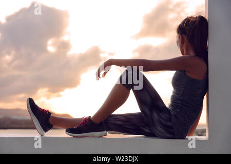 Fitness athlete sports woman relaxing after workout sitting on window looking at sunset view. Silhouette of runner in activewear resting at home on outdoor terrace in sky background.